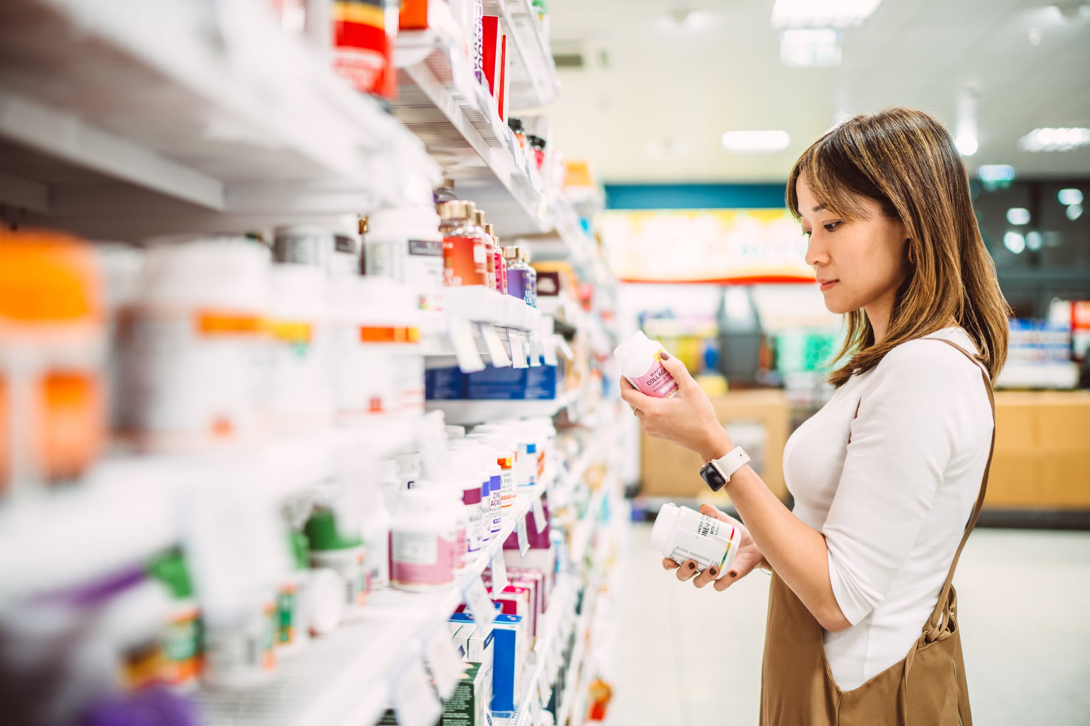 Asian woman examining bottles of vitamins and dietary supplements in a pharmacy or supermarket aisle. She is comparing labels and making a decision on health and wellness products. Concept of healthy lifestyle, nutrition, self-care, and wellness.