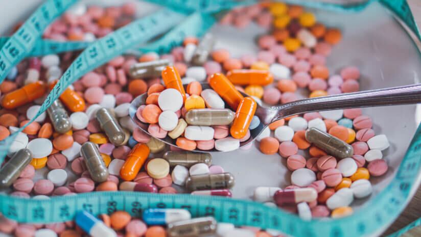 A measuring tape surrounding a spoon filled with supplements surrounded by capsules and pills.