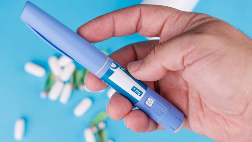 Closeup of hand holding semaglutide pens beside pill bottle and scattered pills on bright blue table, symbolizing healthcare and weight loss support